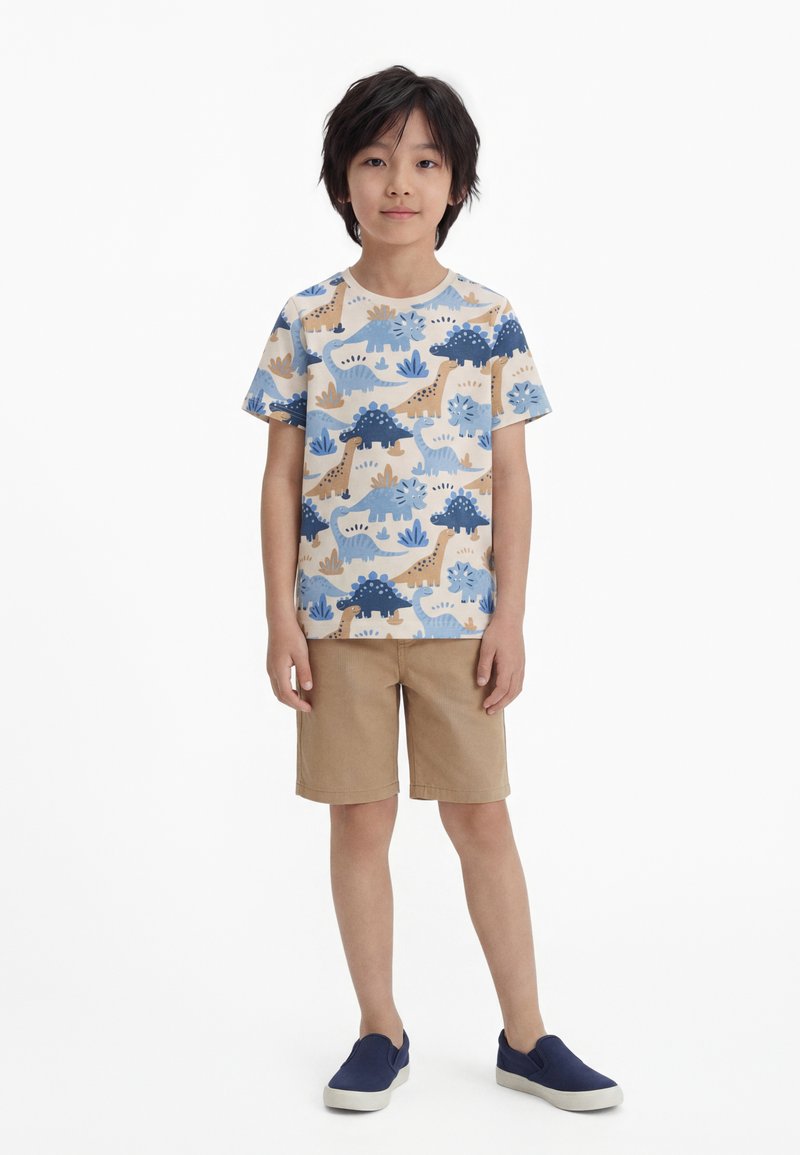Fiú bézs, dinoszaurusz mintás pólóban, világosbarna rövidnadrágban és tengerészkék színű, könnyen felhúzható cipőben, fehér háttér előtt állva.