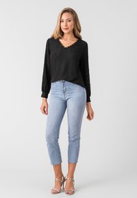 Schwarz langärmliges Oberteil mit V-Ausschnitt, strukturiertem Muster und Spitzenakzenten. Kombiniert mit hellblauen, figurbetonten Jeans und silbernen Riemchensandalen.