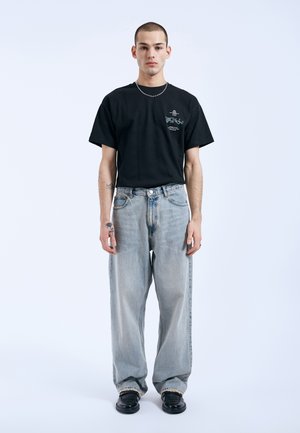 Giovane uomo con capelli corti che indossa una t-shirt nera con grafica, jeans larghi azzurri chiari, scarpe nere e una collana con catenina d'argento in piedi contro uno sfondo bianco.