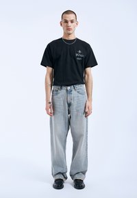 Camiseta de algodón negra con un diseño gráfico, combinada con jeans de mezclilla azul claro de corte relajado y ligero desgaste, y zapatos negros.