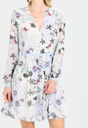 Kvinde iført en lys kjole med blomstermønster, lange ærmer, V-halsudskæring og et lyseblåt bælte i taljen.