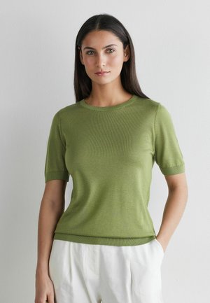 Vrouw met lang donker haar, gekleed in een olijfgroen kortemouwentrui en witte broek, staand tegen een effen lichte muur.