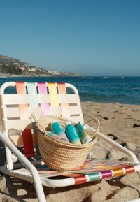 Leżak plażowy z wielokolorowymi paskami trzyma wiklinowy kosz wypełniony butelkami z kremem przeciwsłonecznym na piaszczystym brzegu, zwrócony w stronę oceanu pod czystym niebem.