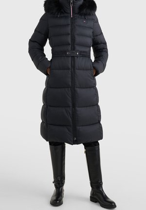 Long manteau d'hiver matelassé noir avec capuche bordée de fourrure, fermeture éclair devant, détail ceinture, porté avec des bottes noires hautes jusqu'au genou.