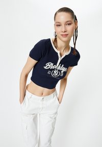 Camiseta corta de color navy con textura acanalada, mangas cortas y tapeta de botones de color crema. Presenta un gráfico de "Brooklyn" en color blanco.
