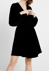 Žena v černých sametových šatech po kolena s dlouhými nabíranými rukávy a uvázaným páskem, stojící na pozadí bílé plochy.