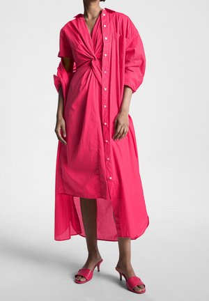 Donna che indossa un vestito midi rosa acceso con davanti annodato e sovrapposizione camicia oversize con bottoni, abbinato a sandali con tacco rosa.