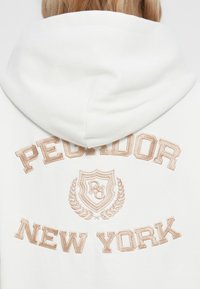 Bijela dukserica s kapuljačom na kojoj su u svijetlosmeđem vezene riječi "PEDLOR" i "NEW YORK", a na leđima je detalj štita i lovora.