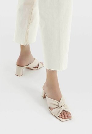 Beige heeled mules with a twisted fabric strap and square toe. The 3-inch block heel provides stability, complemented by a smooth texture.