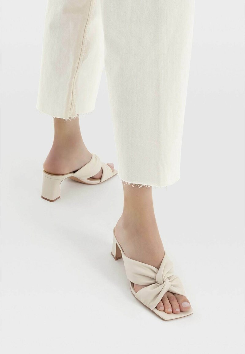 Mules beiges à talons avec une sangle en tissu torsadé et un bout carré. Le talon bloc de 3 pouces offre de la stabilité, complété par une texture lisse.