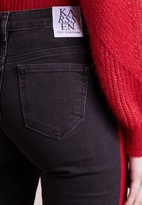 Svarta jeans med en texturerad tyg, med en lapplomme och en märkesetikett på midjebandet. Röda vertikala sidostripes detaljer.