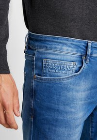 Blå denimjeans med en falmet overflade, der har et baglomme-mærke med teksten "CARS JEANS." Der er en metalknaplukning i taljen.