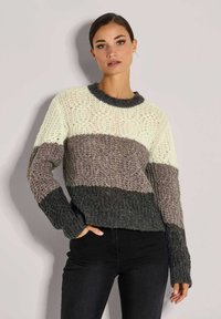 Frau, die einen strukturierten Strickpullover mit breiten horizontalen Streifen in Creme, Taupe und Dunkelgrau trägt, kombiniert mit schwarzen Hosen und vor einer Wand steht.