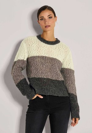 Frau, die einen strukturierten Strickpullover mit breiten horizontalen Streifen in Creme, Taupe und Dunkelgrau trägt, kombiniert mit schwarzen Hosen und vor einer Wand steht.
