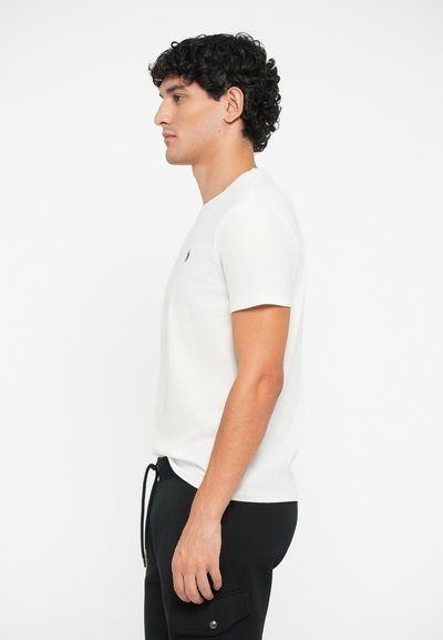 T-shirt branca de manga curta com um decote redondo, apresentando um pequeno logotipo azul no lado esquerdo do peito, combinada com calças pretas.