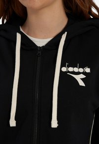 Felpa con cappuccio nera con zip e coulisse color crema, caratterizzata da un logo bianco sul lato sinistro. Realizzata in tessuto morbido con una texture liscia.