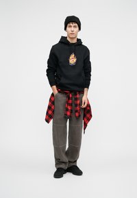 Μαύρη κουκούλα με γραφικά φλόγας και λογότυπο "Santa Cruz", γκρι φαρδύ παντελόνι, κόκκινο και μαύρο καρό φανελάκι δεμένο στη μέση, μαύρα παπούτσια.