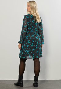 Schwarzes Kleid mit türkisem Blumenmuster, transparentem Material, langen Ärmeln, geradem Schnitt und leicht geschwunghem Saum. Getragen mit schwarzen Ankle Boots.