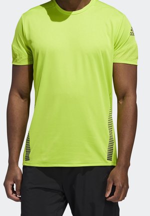 Mann trägt ein hellgelbes kurzärmeliges Sportshirt mit schwarzen Streifendetails und schwarze Shorts, steht vor einem einfarbigen Hintergrund.