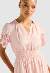 Rochie roz cu mâneci scurte bufante, ornata cu broderie florală. Are un decolteu în V și o talie elastică, rezultând într-o siluetă fluidă.