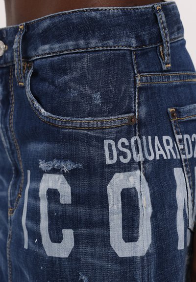 Blaue Jeans aus Denim mit used-Effekten, die eine verblasste Textur, große weiße Buchstaben und metallische Beschläge an der Taille und in den Taschen aufweisen.
