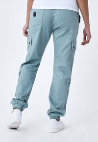 Lichtblauwe cargobroek met elastische boorden, meerdere zijzakken en een zwart logo-detail op de achterkant. Witte sneakers maken de outfit compleet.