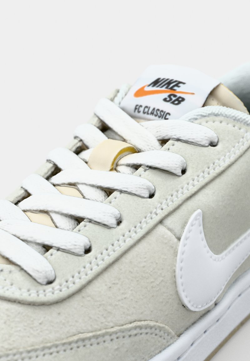 Zapatillas Nike SB Classic en suede gris claro con cordones blancos y suela de goma blanca. Presenta un logotipo de Nike destacado en la lengüeta.