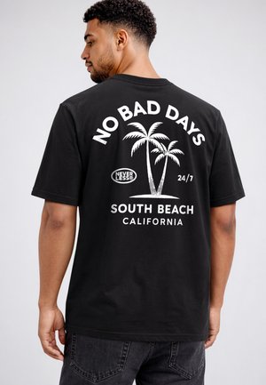 Man wearing black T-shirt with white palm tree graphic and text "No Bad Days, Nevertheless, 24/7, South Beach California" on back.