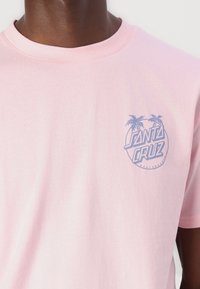 Rožinė medvilnin�ė t-shirt marškinėliai su mažu mėlynu piešiniu, kuriame pavaizduoti palmės ir tekstas "SANTA CRUZ" kairėje krūtinės pusėje. Klasikinio apvalaus kaklo dizainas.