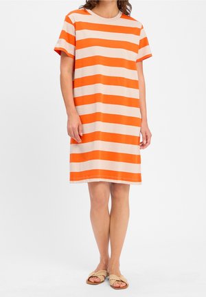 Kobieta ubrana w sięgającą kolan sukienkę T-shirt z poziomymi pomarańczowo-beżowymi paskami oraz beżowe plecione sandały, stojąca na białym tle.