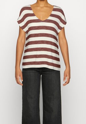 Femme portant une chemise blanche à manches courtes avec de larges rayures horizontales marron et un pantalon noir à jambes larges, debout devant un fond uni.