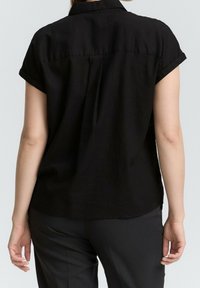 Camicetta nera a maniche corte con colletto, dettaglio delle pieghe posteriori e polsini arrotolati. Realizzata in tessuto leggero, abbinata a pantaloni scuri.