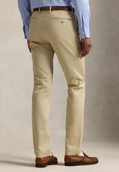 Man draagt beige broek, bruine riem, blauw overhemd met knopen en bruine leren schoenen, staand op een neutrale achtergrond.