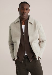 Jachetă gri deschis din amestec de lână, cu guler, închidere cu fermoar, buzunare laterale, purtată peste un pulover maro închis cu fermoar, asortată cu pantaloni închiși la culoare.