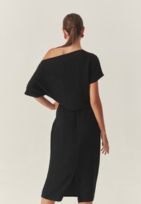 Rochie neagră tricotată, cu umeri descoperiți, mâneci scurte, lungime până la din mediul gambei, țesătură texturată și o deschidere pe spate pentru confortul mișcării.