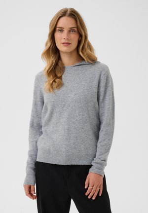 Femme aux longs cheveux blonds ondulés portant un pull à capuche gris clair et un pantalon noir sur un fond clair uni.