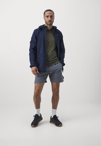 Jūras spēku vieglais veste ar kapuci, olīvu zaļš T-krekls, pelēki šorti, melni sporta apavi ar gumijas zolem un baltas sporta zeķes.