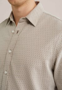 Cămașă de culoare maro deschis, cu un model geometric, guler cu butoni și butoane albe. Are o textură moale și o croială clasică.