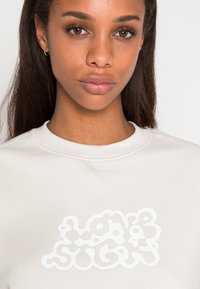 Sweatshirt bege claro com um design gráfico branco texturizado apresentando formas fluidas e curvas na frente, gola redonda e tecido macio.