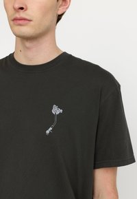 Camiseta de algodón verde oscuro con mangas cortas y cuello redondo acanalado. Presenta un pequeño diseño gráfico blanco en el área del pecho izquierdo.