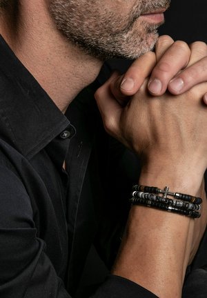 Bracelet noir en perles avec des accents argentés et un charm croix, superposé avec des bracelets supplémentaires plus petits aux textures et couleurs variées.