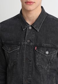 Schwarze Jeansjacke mit Kragen, sichtbaren Metallknöpfen, zwei Brusttaschen und einem kleinen roten Logo-Tag an der linken Tasche.