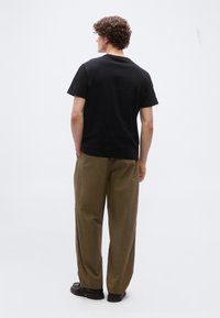 Άντρας με σγουρά μαλλιά, που φοράει μαύρο μπλουζάκι, φαρδιά καφέ παντελόνια και μαύρα παπούτσια, στέκεται με τα χέρια στις τσέπες και κοιτάζοντας προς τα πίσω.