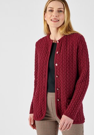 Cardigan in magenta lavorato a maglia con un motivo a treccia testurato, collo rotondo, chiusura con bottoni e due tasche frontali. Indossato sopra una maglia nera e pantaloni a scacchi.