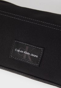 Clutch in tessuto nero con patch del logo Calvin Klein Jeans cucito, dotato di una texture sottile e di cuciture grigie a contrasto.