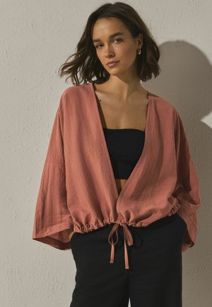 Žena s krátkymi hnedými vlasmi má na sebe voľnú ružovú bundu cez čierny tubový top a čierne nohavice, stojí pred textúrovanou béžovou stenou.