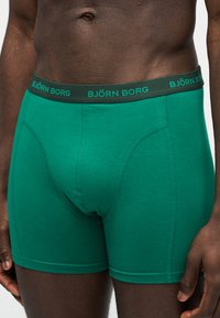 Άνδρας με σκουρόχρωμο δέρμα, κορμός ντυμένος με πράσινα εσώρουχα μποξεράκια Björn Borg με μαύρη μέση, που στέκεται μπροστά σε απλό ανοιχτό φόντο.