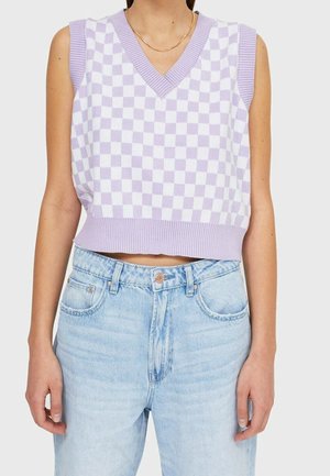 Vrouw draagt een mouwloos paars-wit geblokt gebreid vest met V-hals en lichtblauwe hooggetailleerde jeans, staande tegen een effen achtergrond.