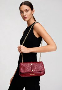 Borsa a tracolla bordeaux con finitura testurizzata, tracolla a catena dorata e dettagli con logo. Forma rettangolare con design a patta pieghevole.