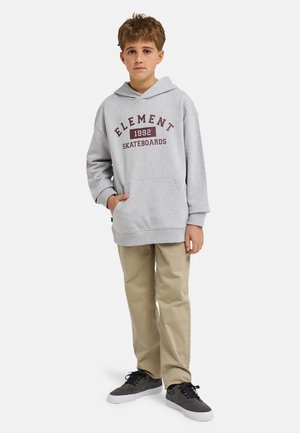 Sweat à capuche gris avec l'inscription "ELEMENT 1992 SKATEBOARDS" en lettres bordeaux, associé à un pantalon beige et des baskets grises sur un fond uni.